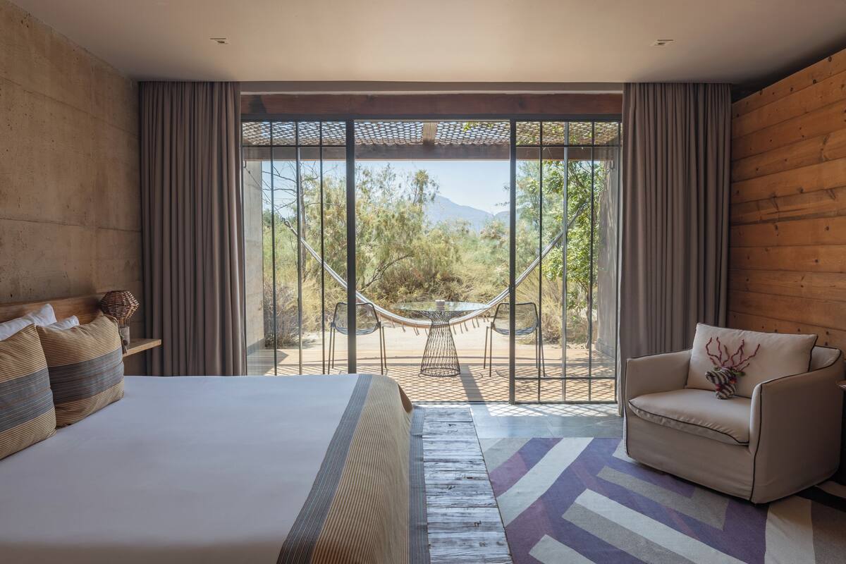 A bedroom with a large bed, a sofa, a dining table with chairs, and a view of the outdoors.