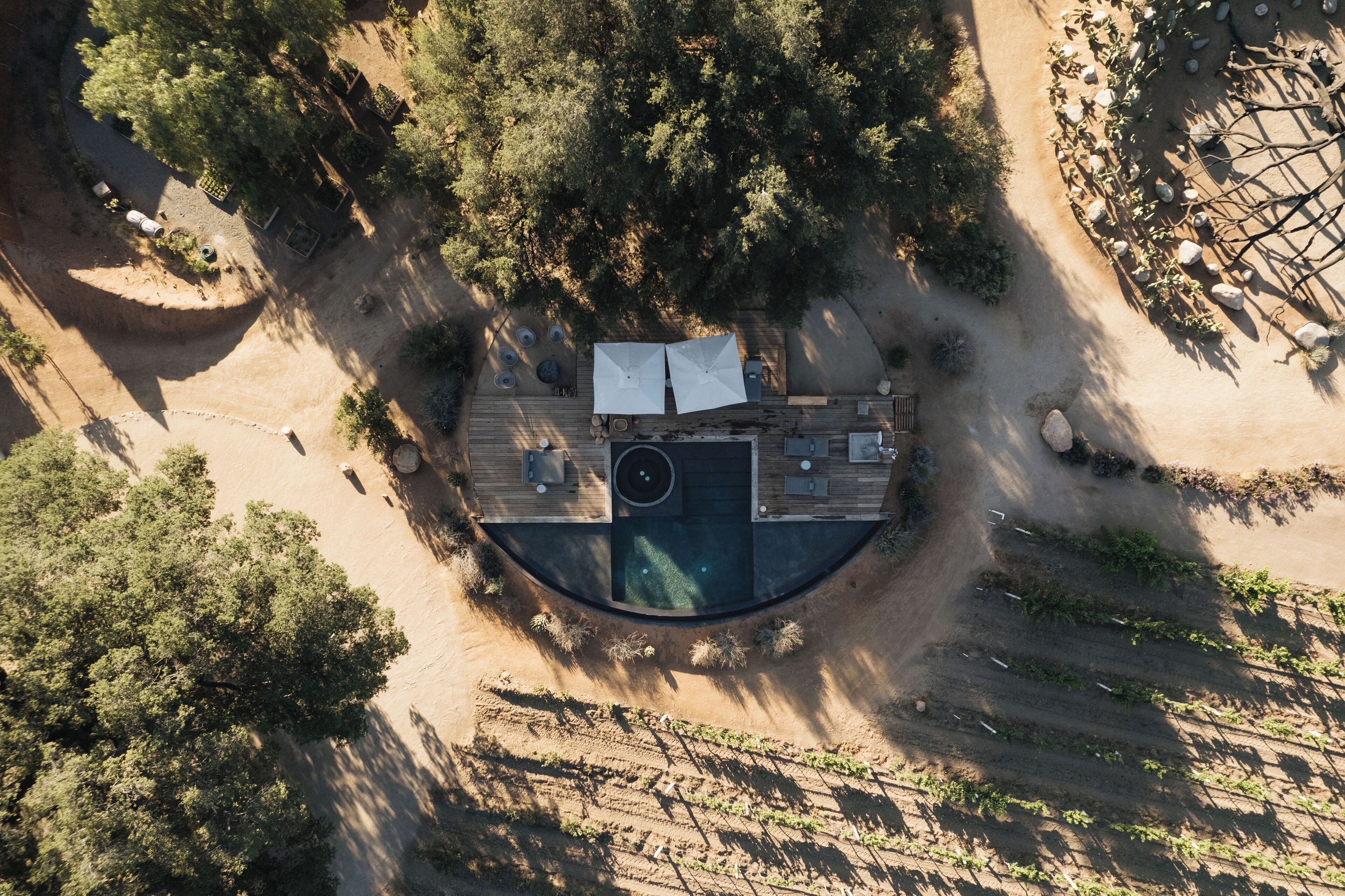 Vista aérea de una terraza de madera circular con piscina, rodeada de árboles y un sendero de tierra.