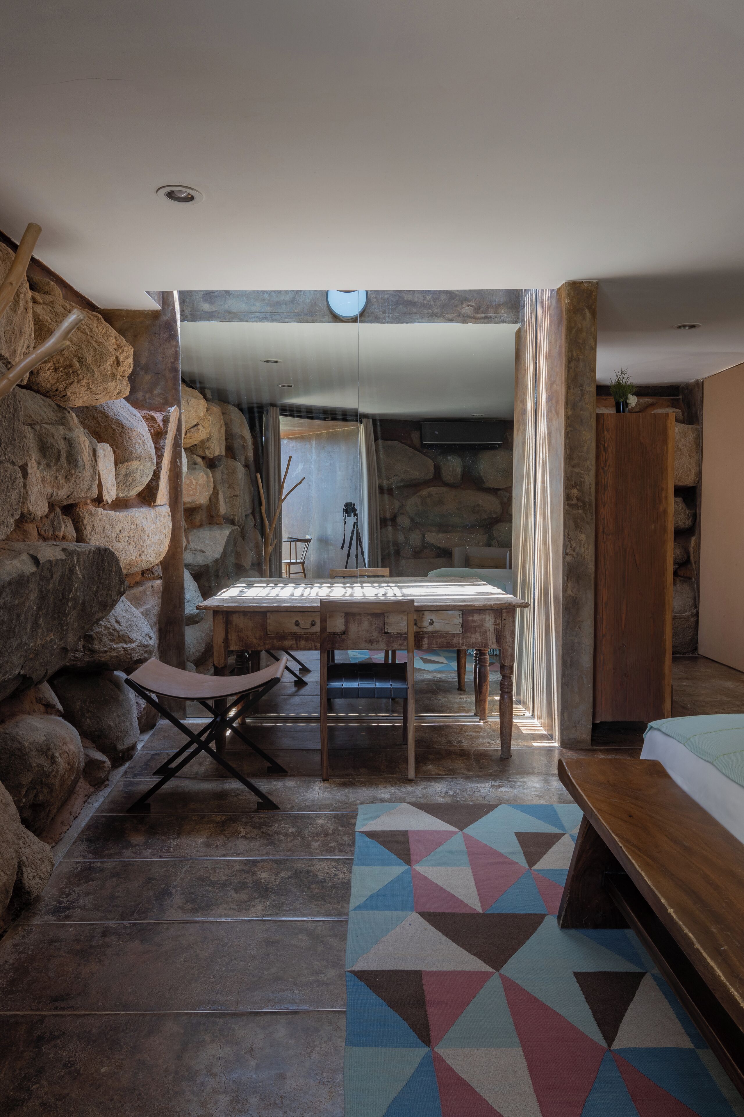 Una habitación con pared de piedra, una mesa de madera, un tapete colorido y una cama.