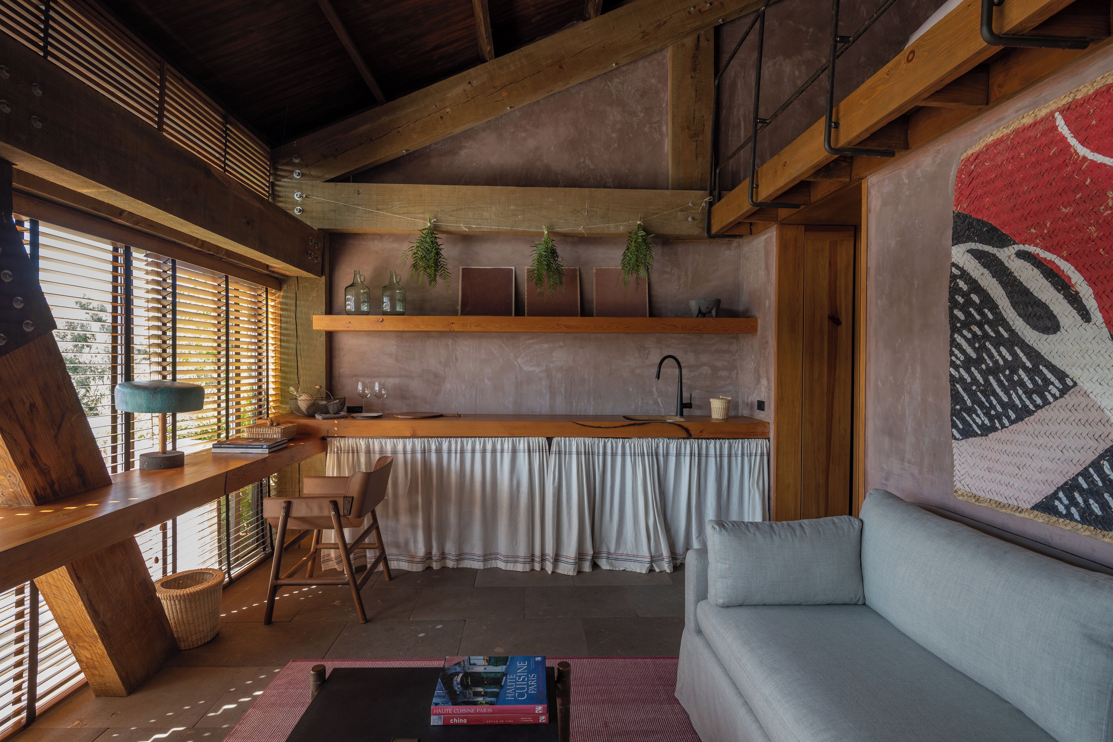 Una sala de estar acogedora con techo de madera, un sofá, una mesa de comedor y una zona de cocina con ventana y plantas.