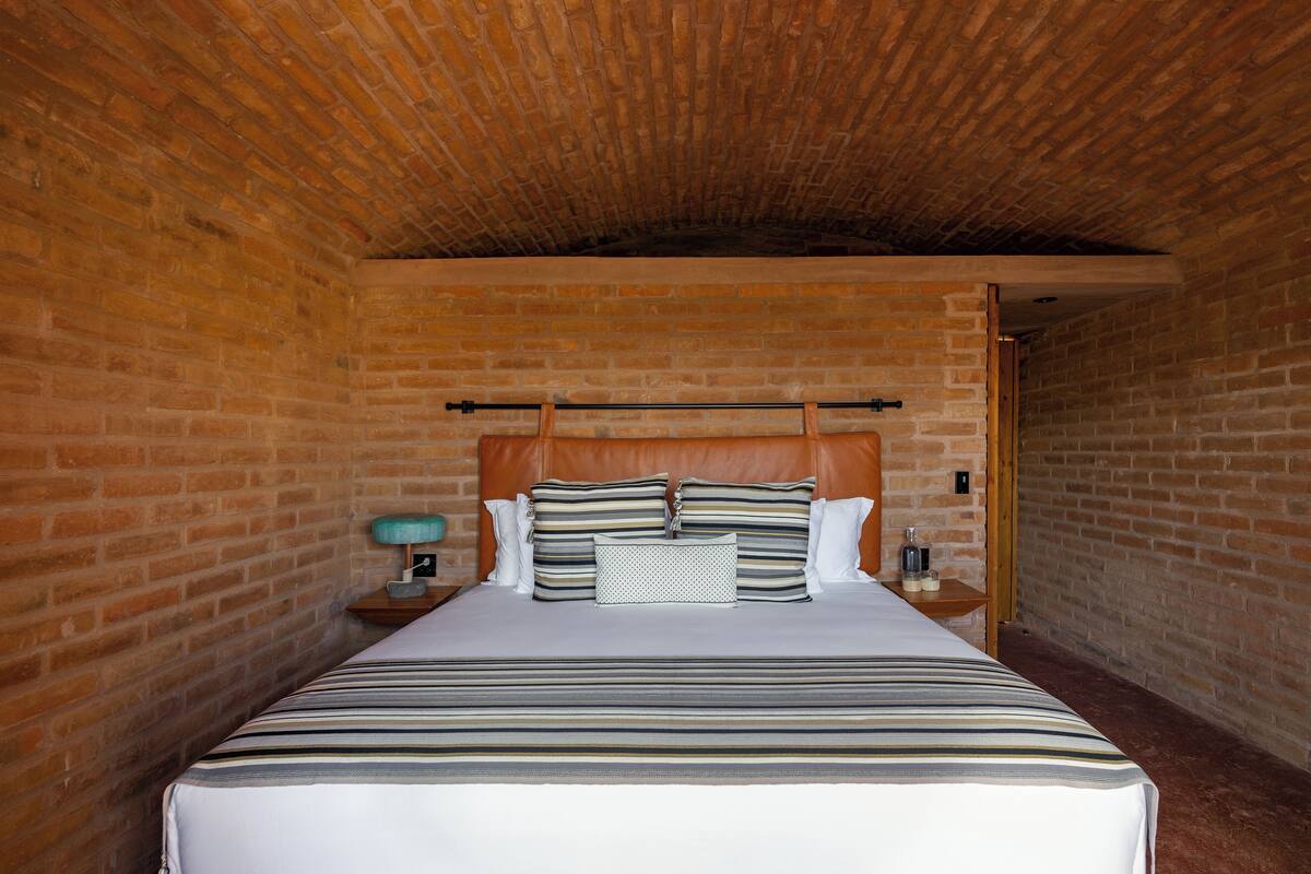 A brick-walled bedroom with a bed, pillows, a nightstand, and a lamp.