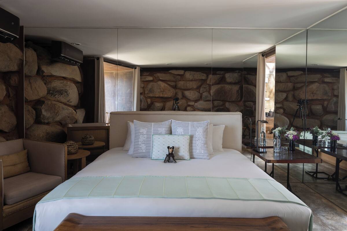 A bedroom with a stone wall, a bed with white bedding, a nightstand, a chair, and a table with a vase of flowers.