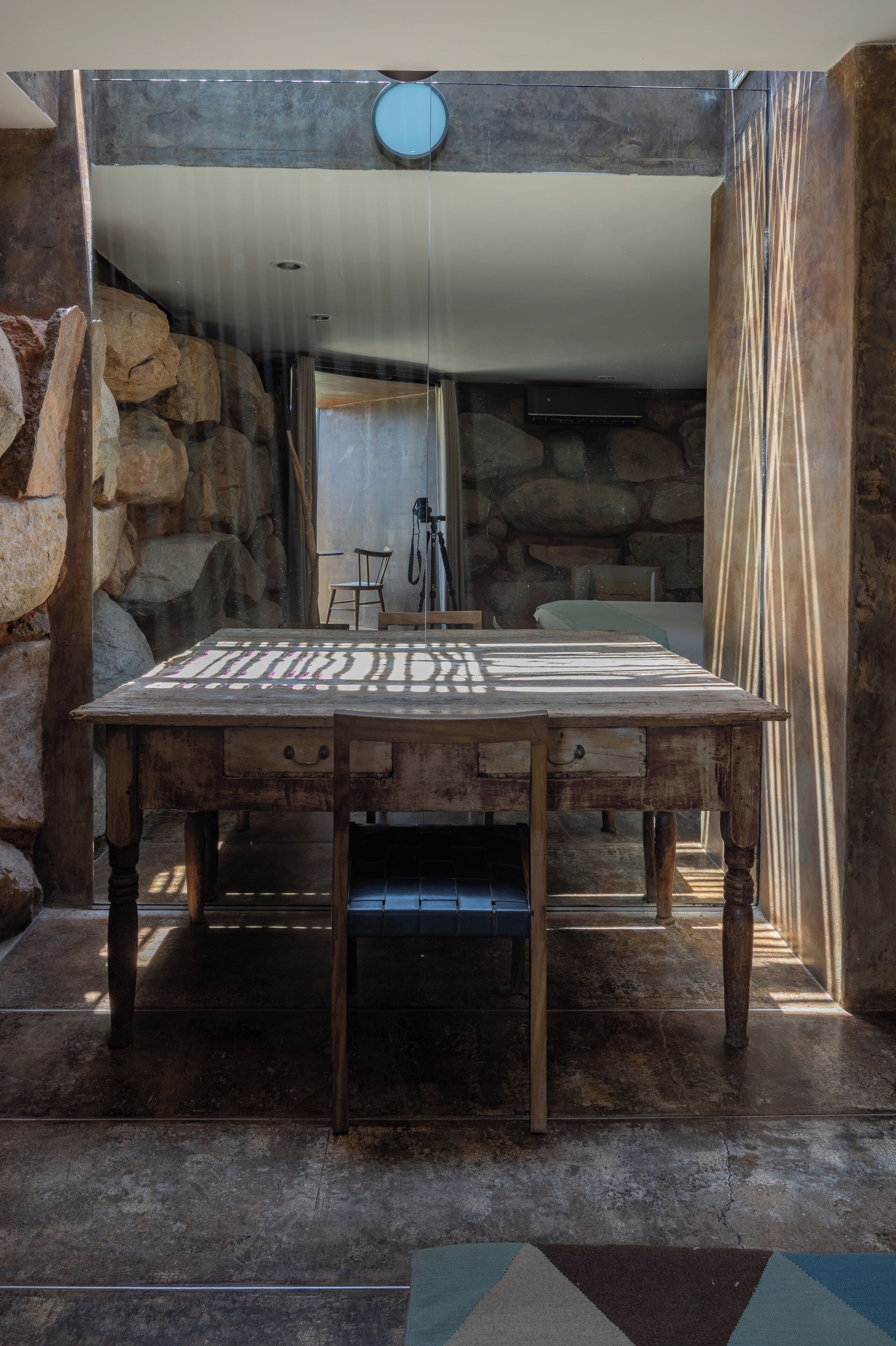Una mesa de madera rústica con mantel estampado, frente a una pared de piedra y un panel de vidrio.