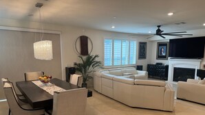 A modern living room with a sectional sofa, a dining table with chairs, a large window with blinds, a fireplace, and a ceiling fan.