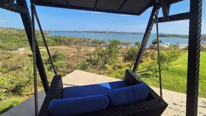 Silla mecedora de mimbre con cojines azules, con vista a un paisaje costero que incluye el mar y colinas.