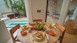 Taula de menjador preparada amb plats, cuplets i una cistella de fruita, a una habitació amb vista a una piscina i zones d'estar a l'exterior.