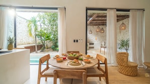 Zona de menjador amb taula rodona preparada per a quatre persones, envoltada de caires de fusta. Hi ha una cadira de vimet i una taula rodona petita amb una sola cadira. L'habitació té una paret de pedra i vista a una piscina i vegetació a l'exterior.