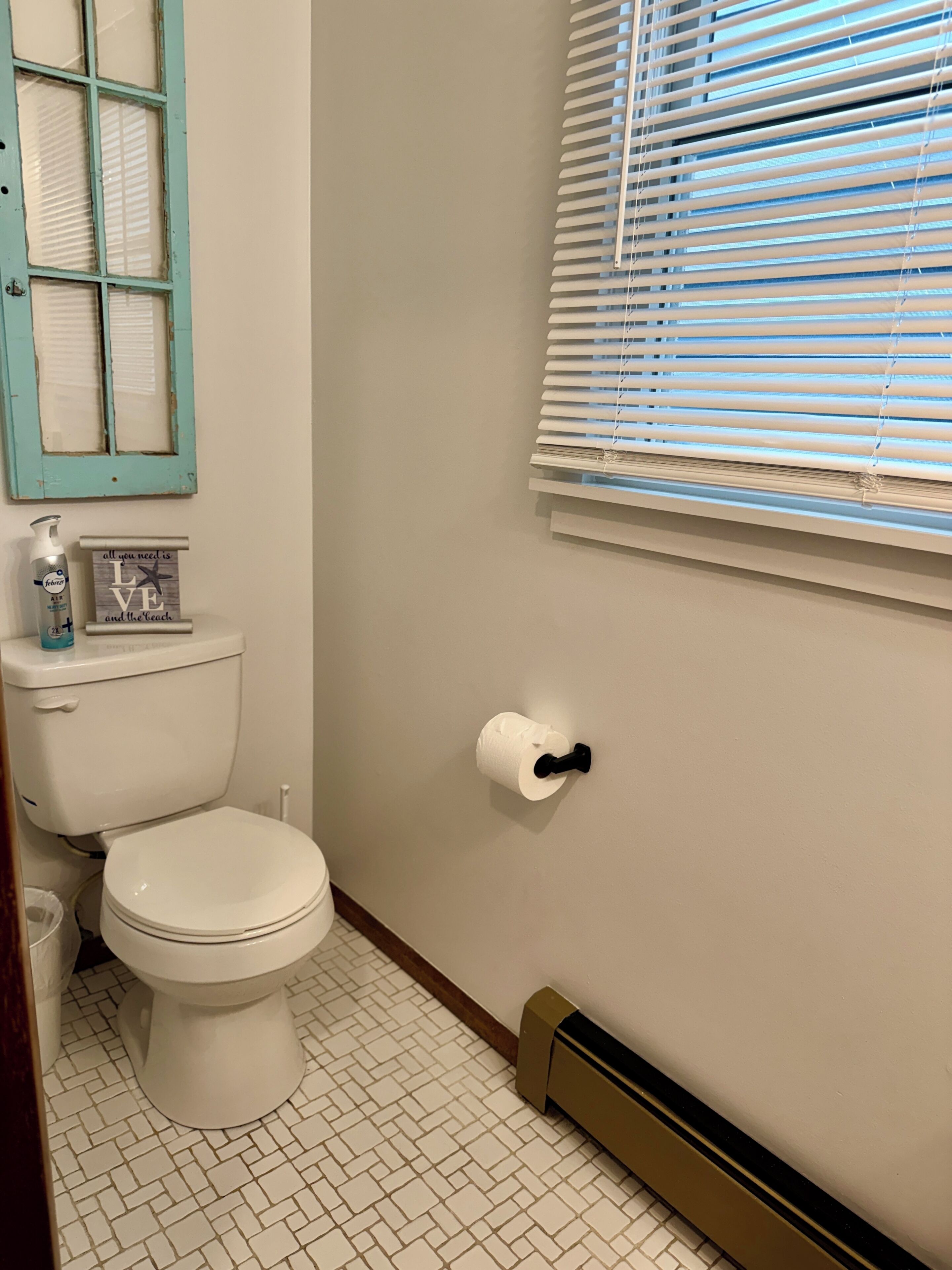 Een badkamer met een toilet, een raam met zonwering, een radiator en een klein plankje met een fles en een bordje.