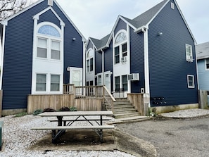 A two-story building with a wooden deck, a picnic table, and a set of stairs leading to the entrance. - Sunny Beach Escape – 2BR Condo Steps from Sand •Badges • Parking (Seaside Heights)