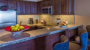 A kitchen with dark wooden cabinets, a stainless steel refrigerator, a microwave, a granite countertop, and a bowl of fruit.