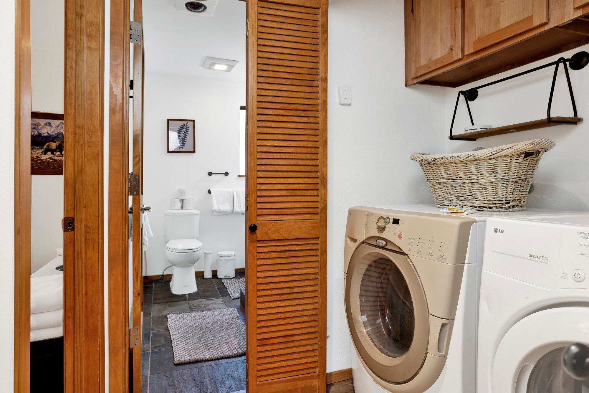 Une buanderie équipée d’un lave-linge, avec un panier posé sur le dessus, et des toilettes visibles à travers une porte en bois.