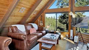 A cozy loft-style room with a leather sofa, a wooden coffee table, and a large window offering a view of trees. - Chimney Peak Chalet#2048 (Big Bear City)
