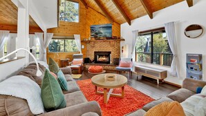 A cozy living room with a fireplace, wooden beams, and large windows. - Chimney Peak Chalet#2048 (Big Bear City)