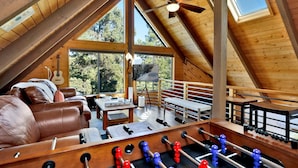 A cozy loft bedroom with a wooden ceiling, a large window, a ceiling fan, a sofa, a table, and a foosball table. - Chimney Peak Chalet#2048 (Big Bear City)