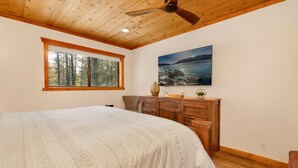 A bedroom with a large bed, wooden headboard, a window with a view of trees, a painting of a lake, and a ceiling fan. - Elmwood Place #2218 (Big Bear City)