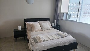Un letto rifatto con cura, con una testiera nera, un comodino con una lampada e una finestra che dà su un edificio.