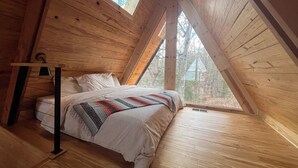 A bedroom with a sloped ceiling, a bed with a patterned blanket, a bedside table, and a window with a view of trees. - Modern lakefront cabin nestled in the piney woods of East Texas. Pet Friendly! (Pittsburg)