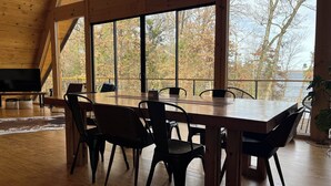 A spacious dining area with a large wooden table and chairs, large windows offering a view of trees and a body of water. - Modern lakefront cabin nestled in the piney woods of East Texas. Pet Friendly! (Pittsburg)