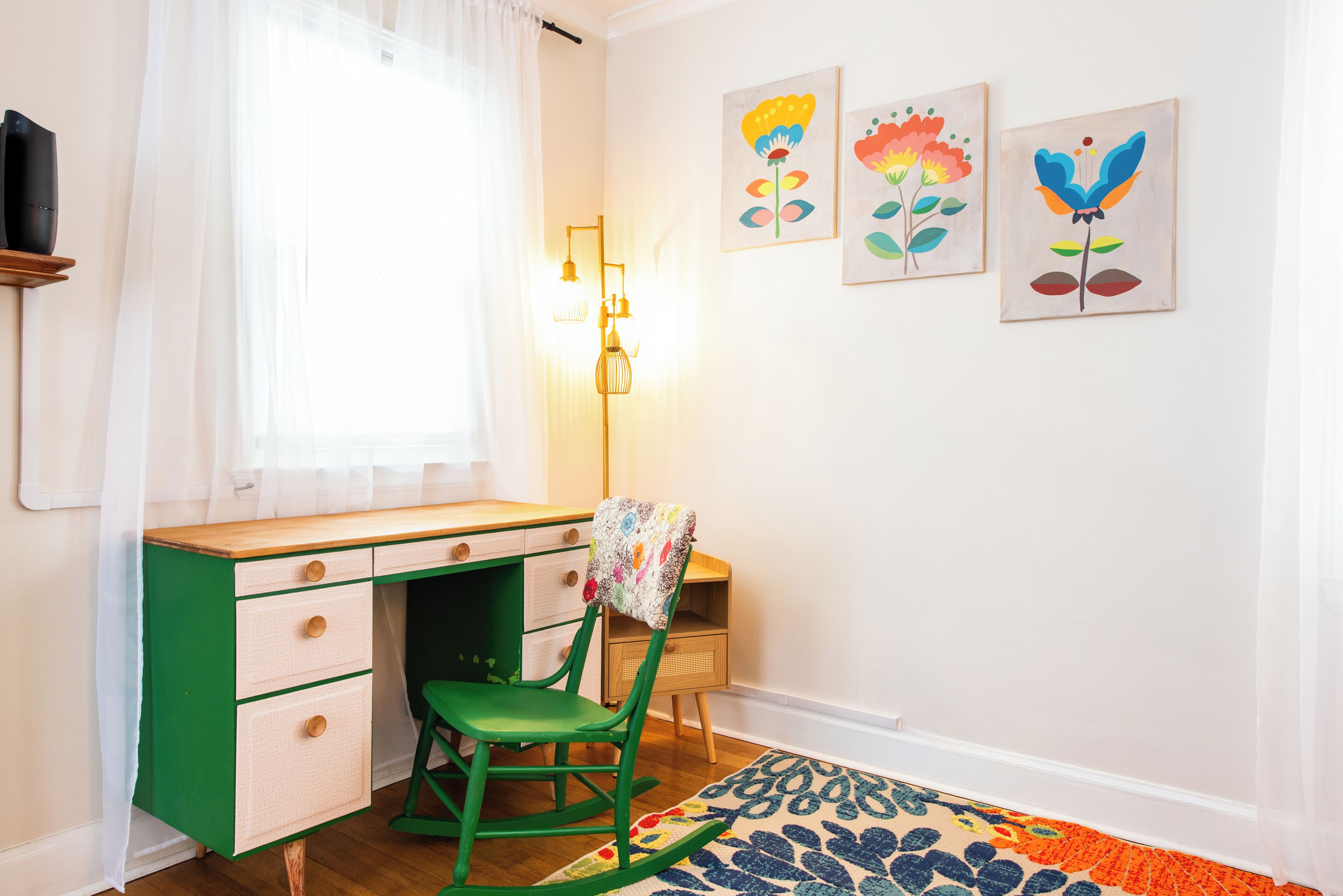 Une pièce avec un bureau vert, un fauteuil à bascule et un tapis coloré. On y trouve trois tableaux accrochés au mur et une lampe.