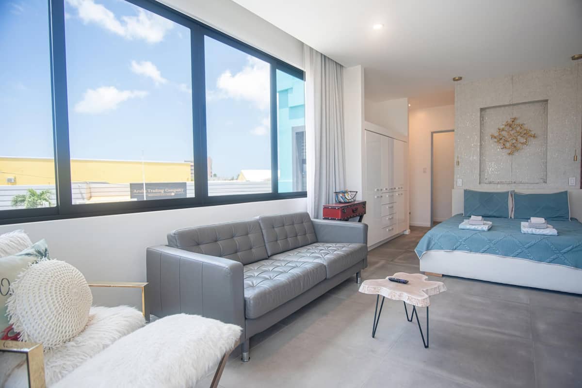 Un dormitorio moderno con un ventanal grande, un sofá gris, una cama con ropa de cama azul y una mesita pequeña.