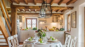 A dining area with a round table set for six, surrounded by white chairs. The room features exposed wooden beams on the ceiling and white cabinetry in the background. - Grumo Farmhouse in Pescaglia (Pescaglia)