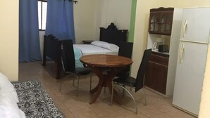 A small, furnished room with a bed, a table with chairs, a refrigerator, and a window with curtains. - Beach Area Studio (Playas del Coco)