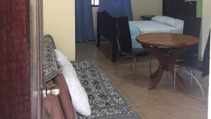 A room with a bed, a table, and a chair. There is a window with blue curtains and an air conditioning unit above it. - Beach Area Studio (Playas del Coco)