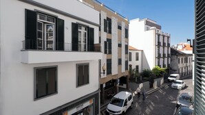 A multi-story building with balconies, a ground-floor shop, and parked cars on a cobblestone street. - In the centre of Funchal - Apartamento do Carmo I (Funchal)