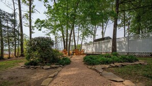 Un sendero de grava que conduce a una terraza de madera con sillas y una mesa, rodeado de árboles y una cerca blanca.
