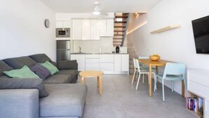 A modern living room with a grey sofa, a small wooden table, and a kitchen area with white cabinets and a built-in oven. - Home2Book Charming Apt Costa Adeje, Pool&Terrace (Playa de las Americas)
