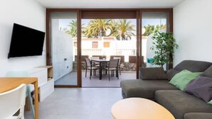 A modern living room with a sofa, a coffee table, and a TV mounted on the wall. There is a sliding glass door leading to an outdoor patio area with a table and chairs. - Home2Book Charming Apt Costa Adeje, Pool&Terrace (Playa de las Americas)