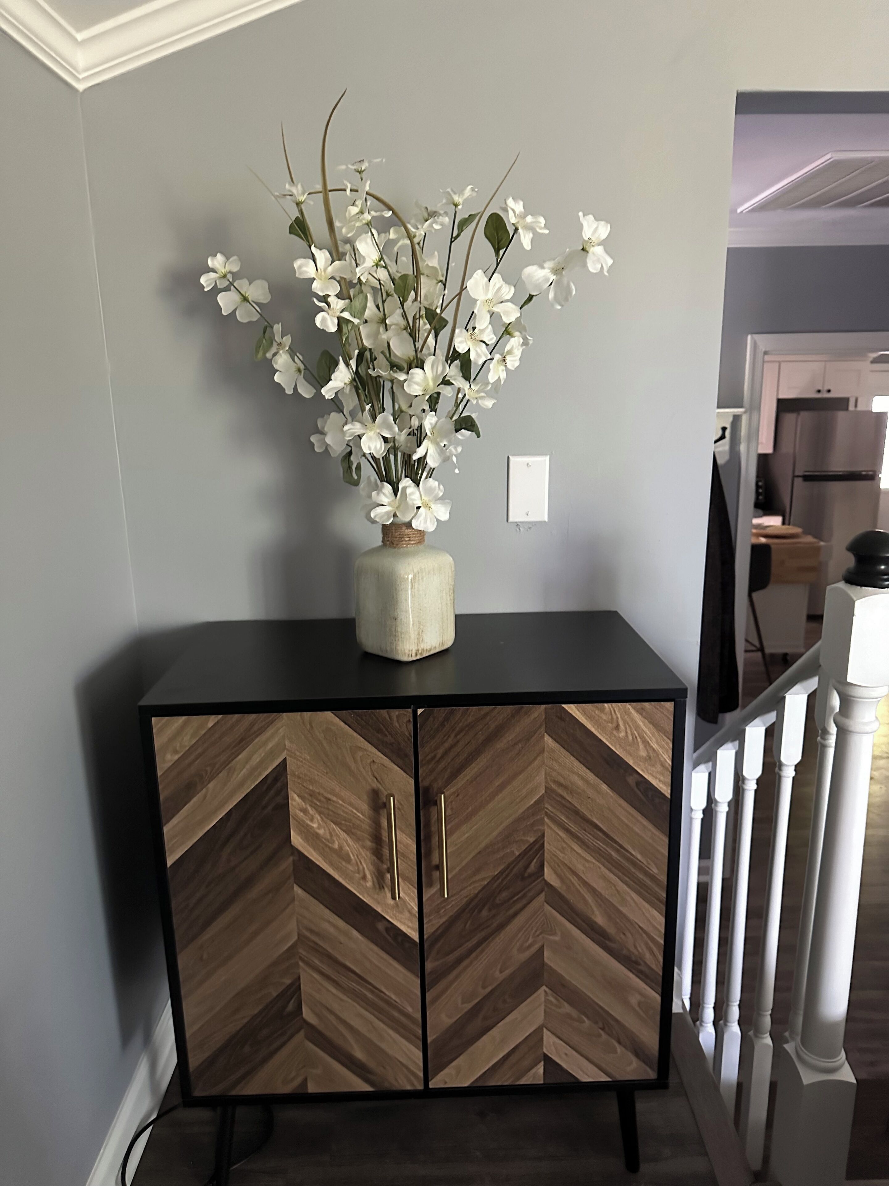 Un meuble en bois à motifs en chevrons, surmonté d’un vase de fleurs blanches, un interrupteur blanc sur le mur et un escalier avec des balustres blanches.