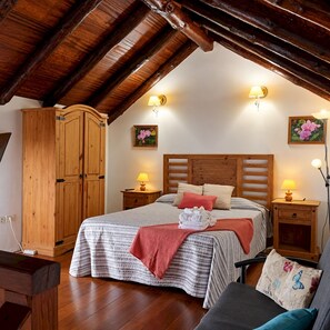 A bedroom with a wooden ceiling, a bed with a striped bedspread, two bedside lamps, a wooden wardrobe, and two wall-mounted flower paintings.