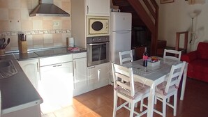 Una cocina compacta con armarios blancos, estufa, horno y refrigerador. En el área de estar contigua, hay una mesa de comedor con sillas y un sofá rojo.