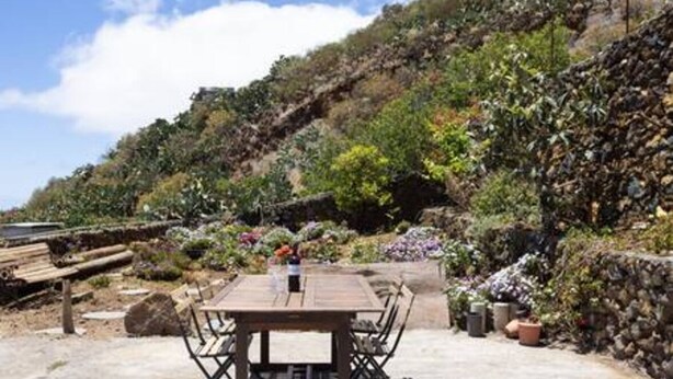 An outdoor patio with a wooden table and chairs, surrounded by a stone wall and lush vegetation. - La Pestilla 3 - Cozy rustic house, El Hierro (Taibique)
