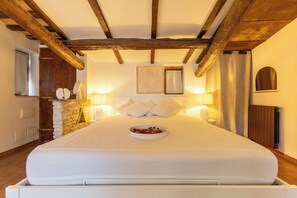 A bedroom with a large bed, wooden beams, a stone wall, a small table with a bowl of fruit, and two wall-mounted lamps.