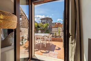 A view through a glass door to an outdoor patio with a table and chairs, potted plants, and a building with a tiled roof in the background.