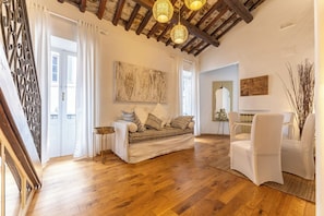 A room with a wooden floor, a sofa with patterned cushions, a white chair, a table with a lamp, and a painting on the wall.