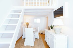 A white interior with a staircase, a dining area with a table and chairs, a lamp, a cabinet, and a television mounted on the wall.