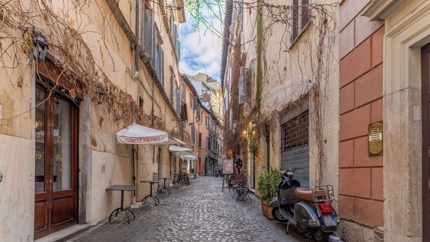 Une rue pavée avec une ruelle étroite, des tables installées à l’extérieur et un scooter garé.