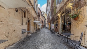 Une rue pavée avec des tables et des chaises installées à l’extérieur, des bâtiments dotés de volets et un ciel dégagé.