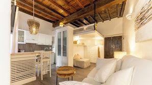 Un intérieur moderne comprenant une cuisine, un coin repas et un salon, avec des poutres en bois, des meubles blancs et un lustre.
