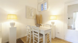 Una mesa de comedor blanca con cuatro sillas, dos lámparas fijadas a la pared, una ventana con contraventanas y un cuadro en la pared.