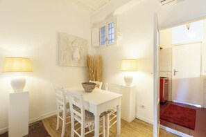 A dining area with a white table, chairs, and two lamps. There is a window with shutters and a painting on the wall.