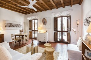 Une pièce avec un plafond en bois, un canapé blanc, une table à manger avec des chaises, une commode blanche et des tabourets en osier.