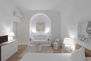 Ein modernes, minimalistisches Schlafzimmer mit einem weißen Sofa, einem Flachbildfernseher, einem Nachttisch mit Lampe und einem Wandkunstwerk.