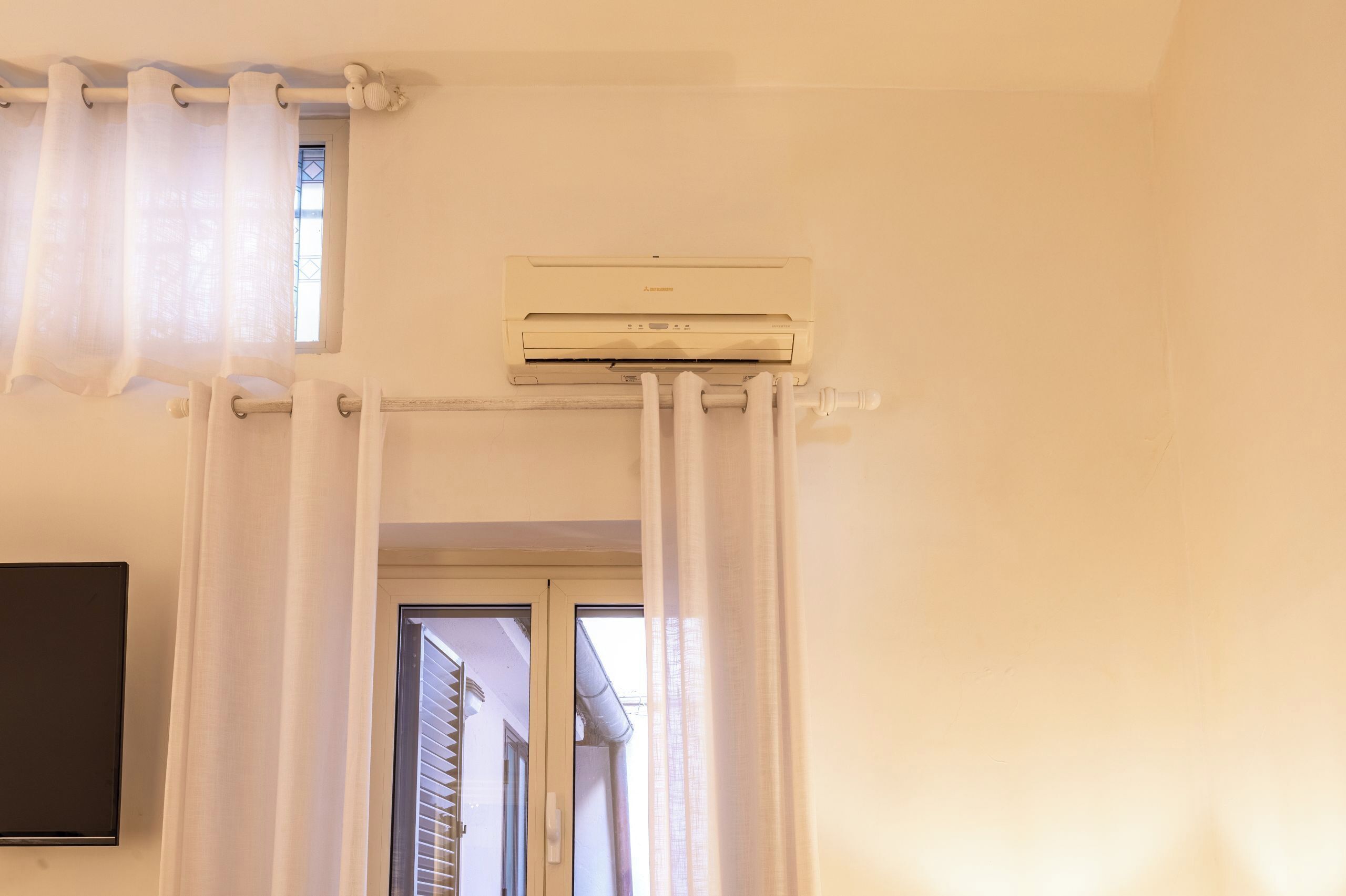 Una habitación con aire acondicionado instalado en el techo, una puerta con vistas al exterior y una barra para cortinas con cortinas blancas.