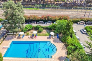 A swimming pool with a geometric tile design, surrounded by lounge chairs and umbrellas, with a parking area and greenery in the background. - Homiday - Residence Prestige, Interno 10 (Pineto)