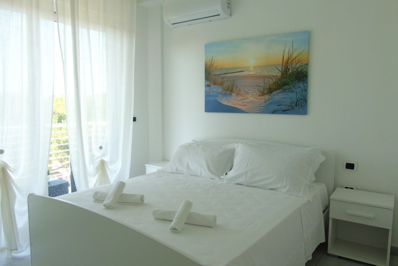 A hotel room with a bed, a nightstand, a painting of a beach scene, and a window with sheer curtains.