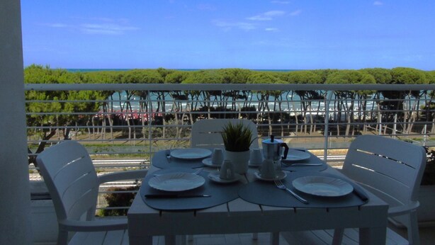 Taula per a quatre amb cadires blanques, una planta en test i vista al mar i vegetació.